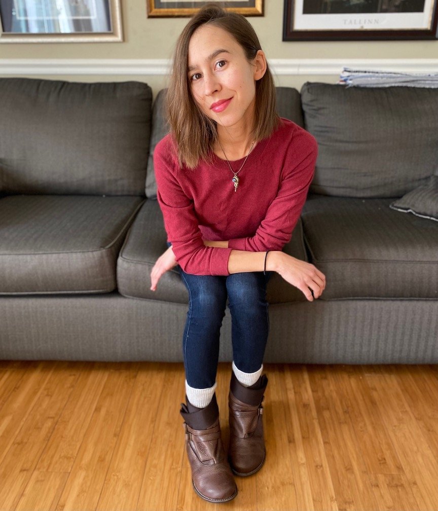 A woman sitting on the couch smiling at the camera wearing a red sweater, jeans, and Lisbeth Joe Boulder Boots in brown leather