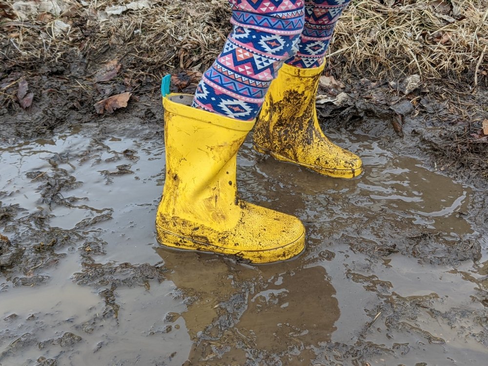 A pair of bright yellow barefoot rain boots from Ten Little being worn by a child in thick mud and water