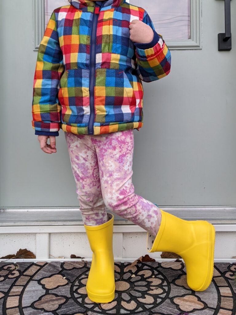 A child wearing a plaid jacket and pink tie dye pants paired with lemon yellow rain boots, an affordable footwear option from Ten Little Kids