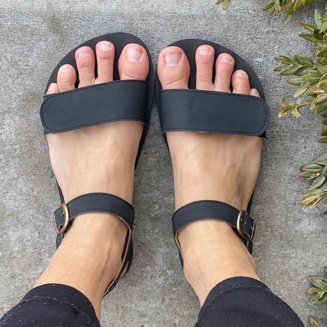 A top down view of a pair of feet standing on concrete wearing black vegan Tikki Shoes barefoot sandals - Vibe model