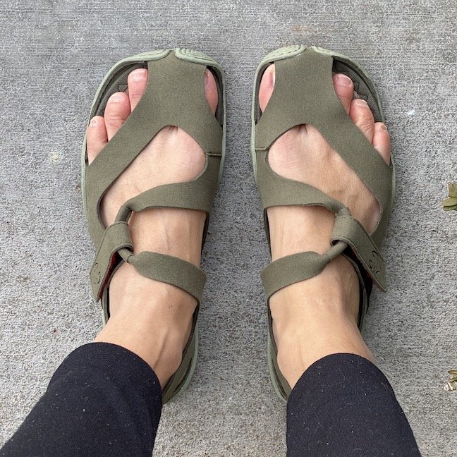 A top down view of a pair of feet standing on concrete wearing Wildling Shoes Forest Feather barefoot sandal made of green microfiber material