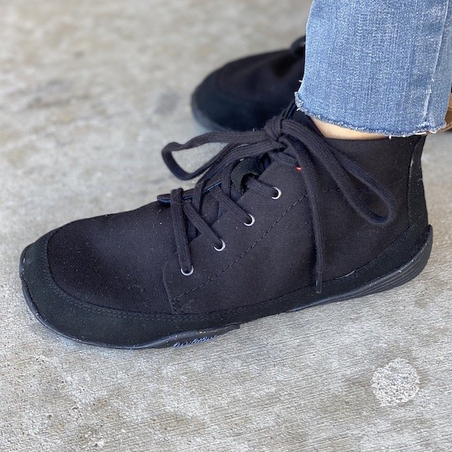 A side view of a foot standing on concrete wearing the Flying Fox black high top barefoot shoes from Wildling