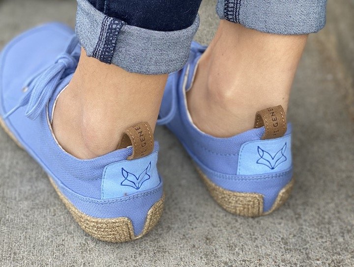 A behind view of a pair of feet standing on concrete wearing Wildling Shoes Kul blue barefoot lace up shoes with the fox logo visible on the heels