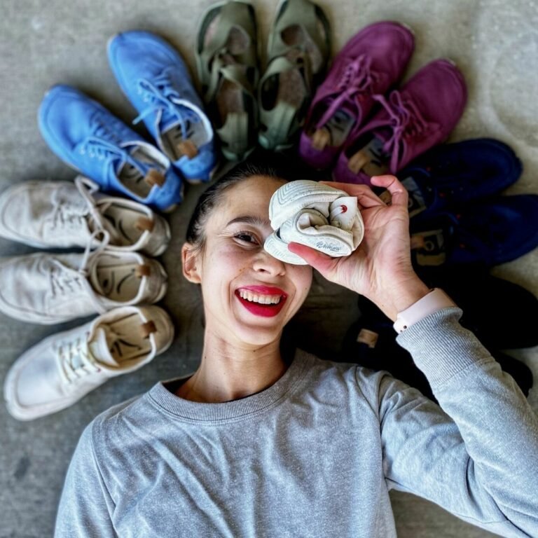 A close up of a woman's face lying on the ground, smilling at the camera with a circle of Wildling Shoes SPring Summer 2022 Collection. She is holding one white Wildling shoe rolled up into a ball over her left eye.