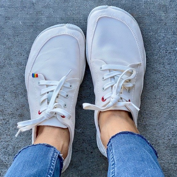 A top down view of a pair of feet standing on concrete wearing the Wildling Shoes Tanuki Niji lightweight white vegan barefoot lace up shoe