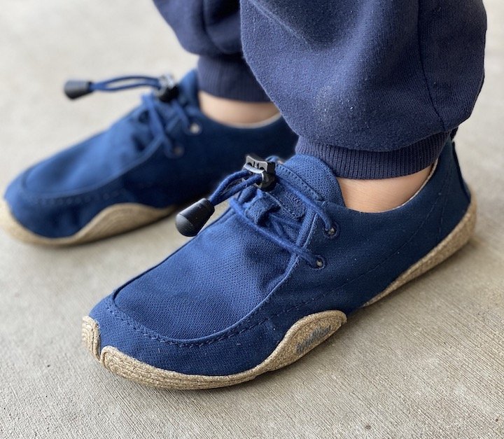 A child's feet wearing blue Wildling Shoes Tengri with elastic laces and the Wildling brand visible on the outsole