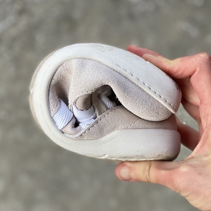 A close up of a hand holding the Xero Shoes Kelso barefoot sneaker in pink leather with a white sole, rolled up into a ball to show its flexibility