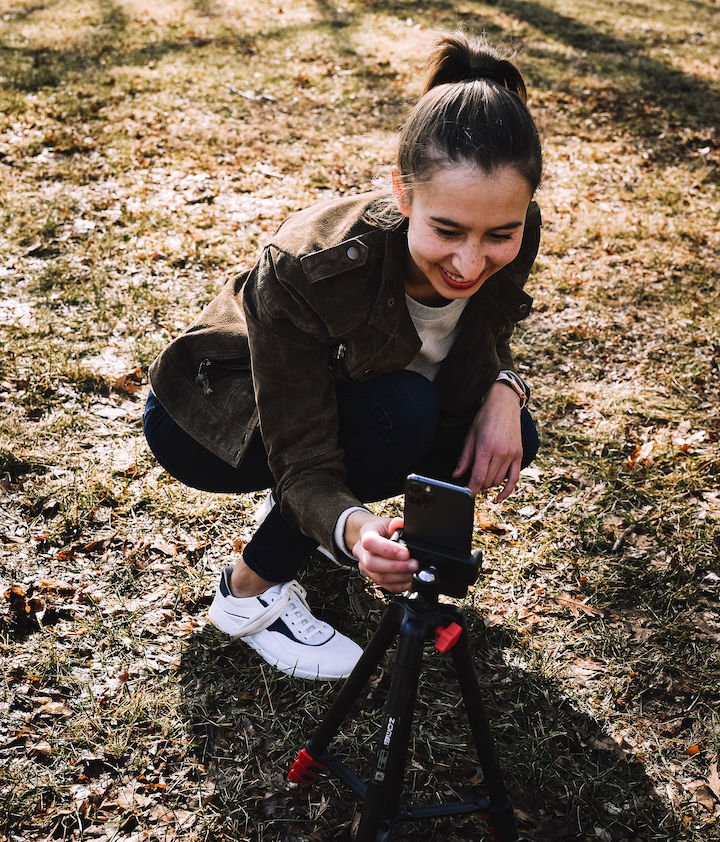 A woman squatting down on grass smiling and looking at a phone on a tripod wearing a suede jacket, jeans, and Groundies Flow Performance barefoot sneakers in white leather