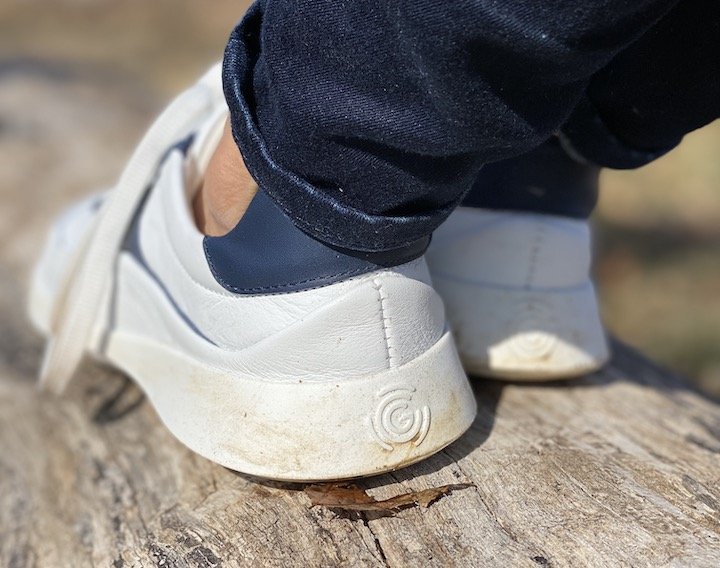 A close up view of a pair of feet wearing white leather Groundies Performance Flow barefoot sneaker on a log with the heel and Groundies logo in focus