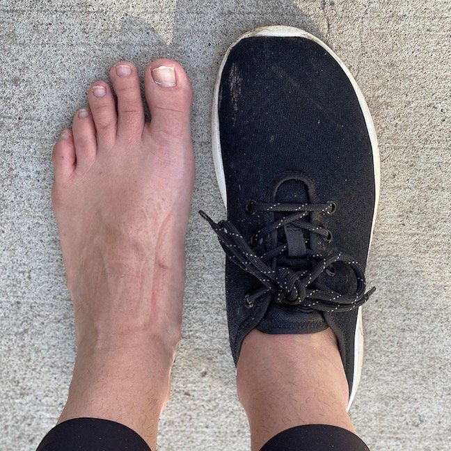 A top down view of a pair of feet standing on concrete with the left foot bare and the right foot wearing a black Groundies Performance Balance barefoot sneaker