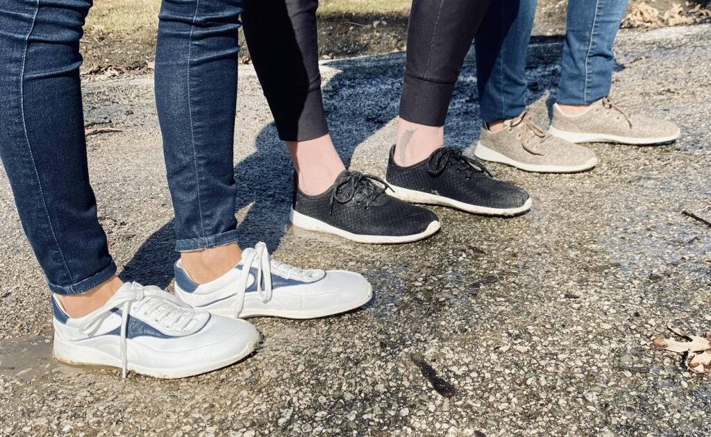 A side view of a row of 3 people's feet from the knees down standing on asphalt outside wearing Groundies Performance barefoot sneakers. In the front is the Flow in white leather, in the middle is Balance in black, and in the back is Relax in taupe wool.