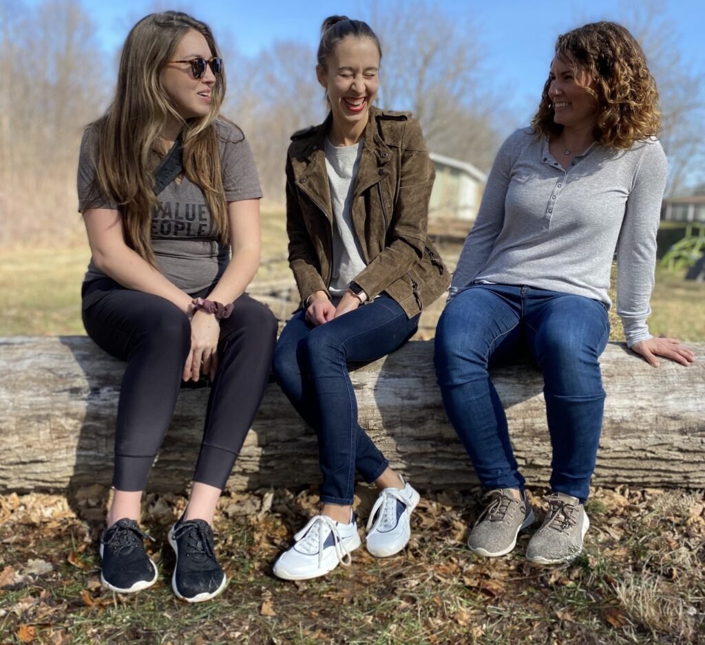 A row of 3 women sitting on a log outside talking and laughing wearing Groundies Performance barefoot sneakers. On the left is the Balance in black, in the middle is Flow in white leather, and on the right is Relax in taupe wool.