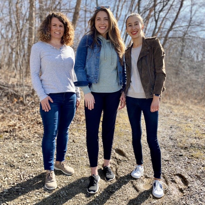 A row of 3 women standing on dirt outside near trees smiling at the camera wearing Groundies Performance barefoot sneakers. On the left is Relax in taupe wool, in the middle is Balance in black, and on the right is Flow in white leather