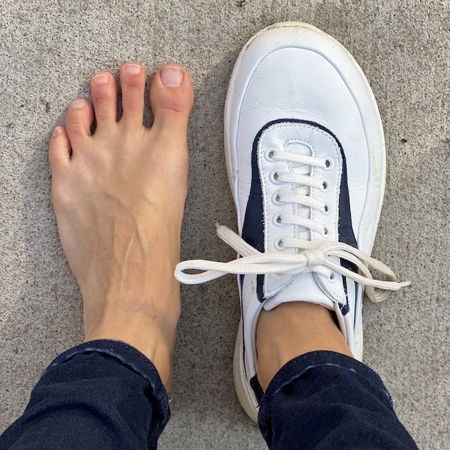 A top down view of a pair of feet standing on concrete with the left foot bare and the right foot wearing a white leather Groundies Performance Flow barefoot sneaker