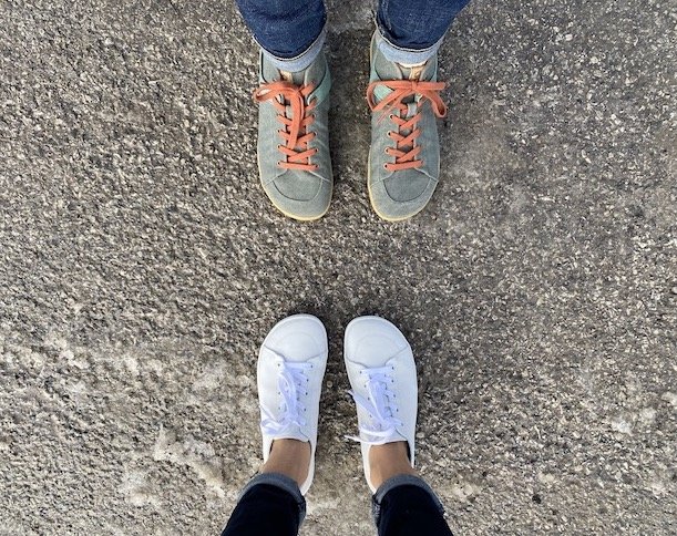 A top down view of two pairs of feet facing each other wearing Mukishoes barefoot shoes. On the top is the Ivy high top sneaker and on bottom is the Raw Leather Cloud in white