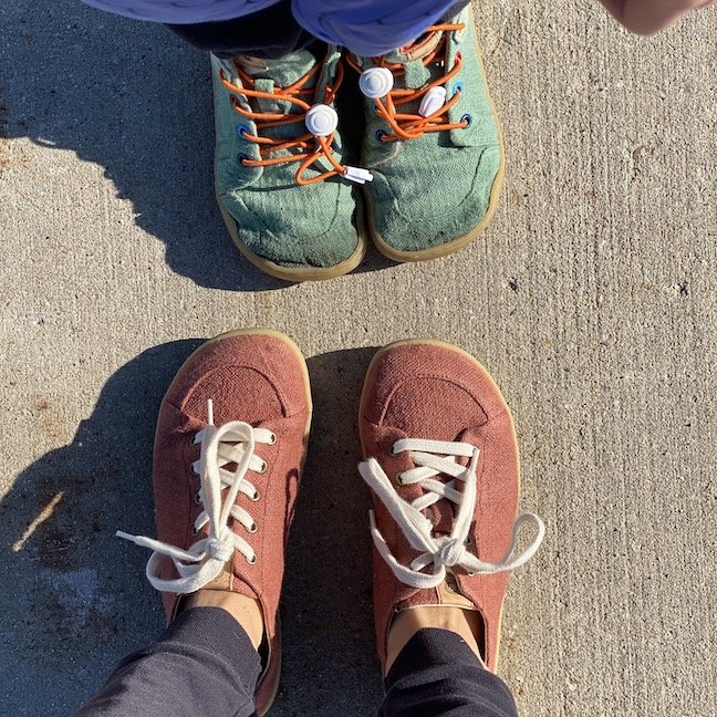 A top down view of two pairs of feet facing each other wearing Mukishoes barefoot shoes - on the top are the feet of a small child wearing Mini Moss green sneakers with orange laces and rainbow eyelets. On bottom is an adult's feet wearing the Saffron rust colored sneaker with white laces