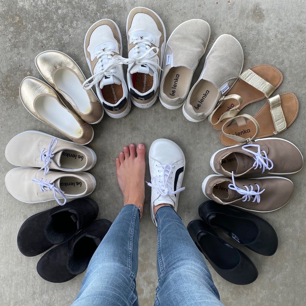 A top down view of a pair of feet standing on concrete with on bare and the other wearing a white Brooklyn sneaker from Be Lenka Barefoot, with a circle of more barefoot shoes from Be Lenka in light spring colors surrounding the feet