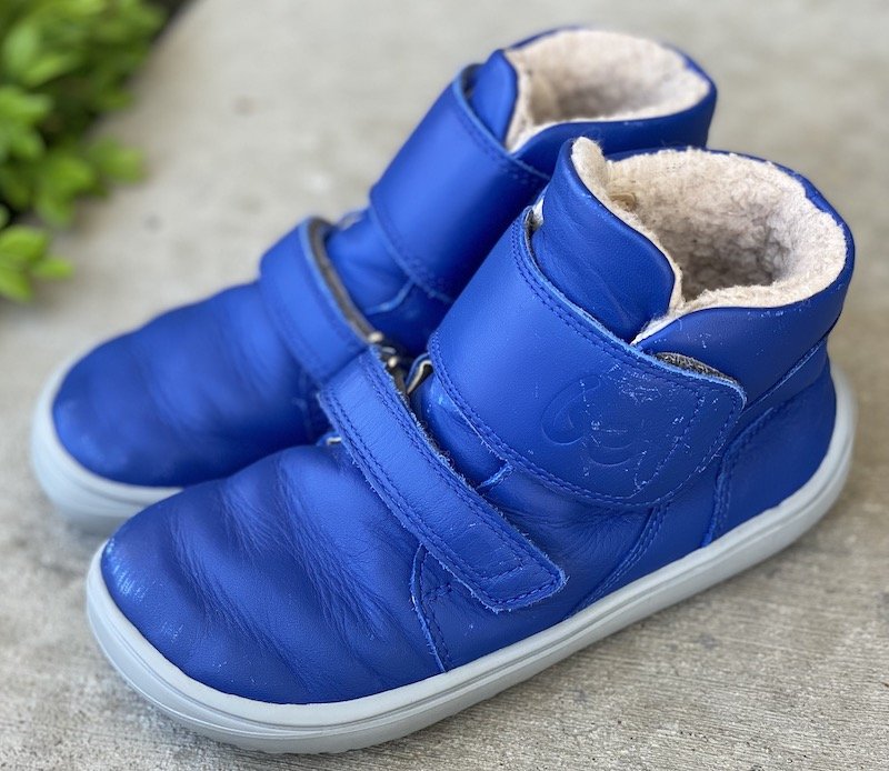 A side close up view of a pair of Be Lenka Panda barefoot winter booties for kids in blue leather sitting on concrete