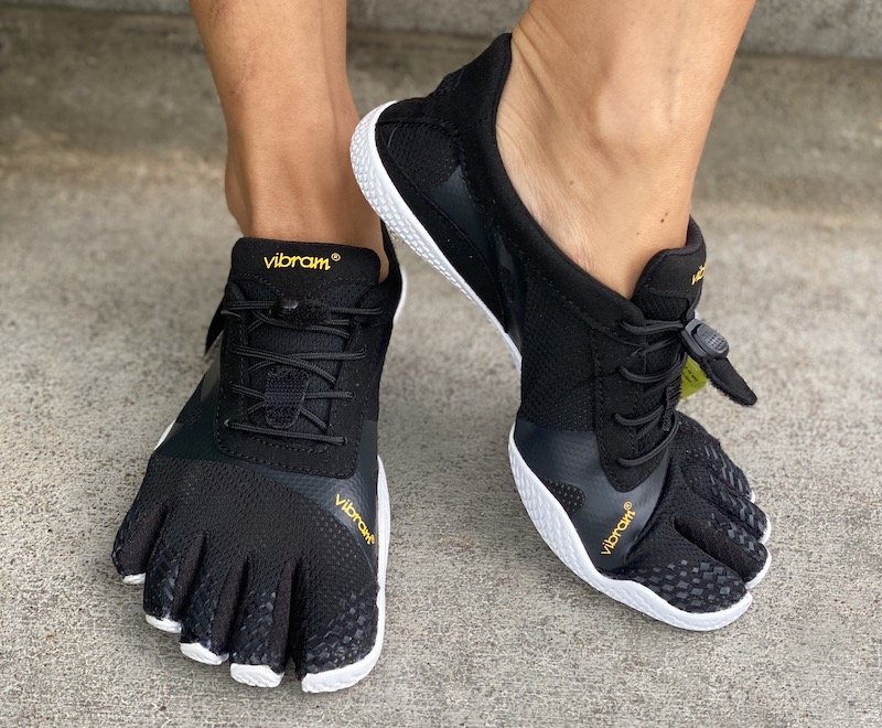 A front view of a pair of feet standing on concrete wearing Vibram Five Fingers KSO barefoot running shoes in black with one foot pointed