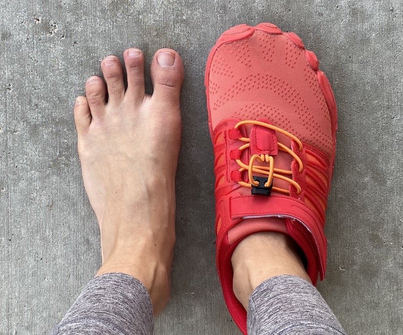 a top down view of a pair of feet standing on concrete. the left foot is bare and the right is wearing an orange Whitin trail barefoot running shoe