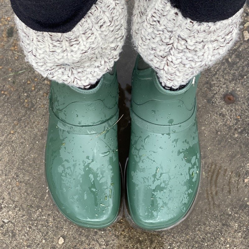 A top down view of a pair of feet wearing green Xero Shoes Gracie rubber barefoot rain boots standing in a puddle