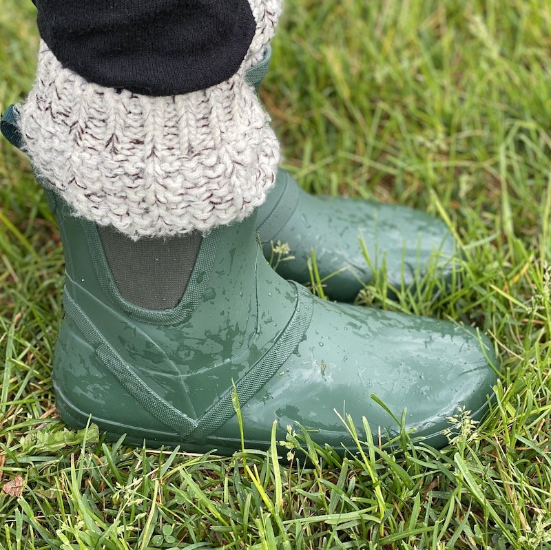 A side view of a pair of feet wearing green Xero Shoes Gracie rubber barefoot rain boots standing in wet grass