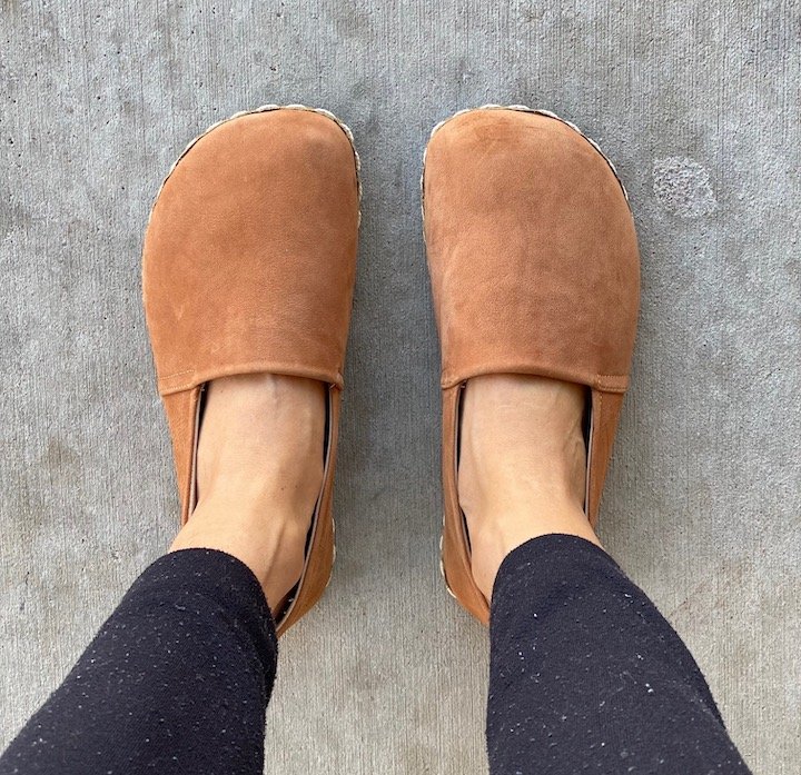 A top down view of a pair of feet standing on concrete wearing Groundx barefoot slip on shoes in clay suede