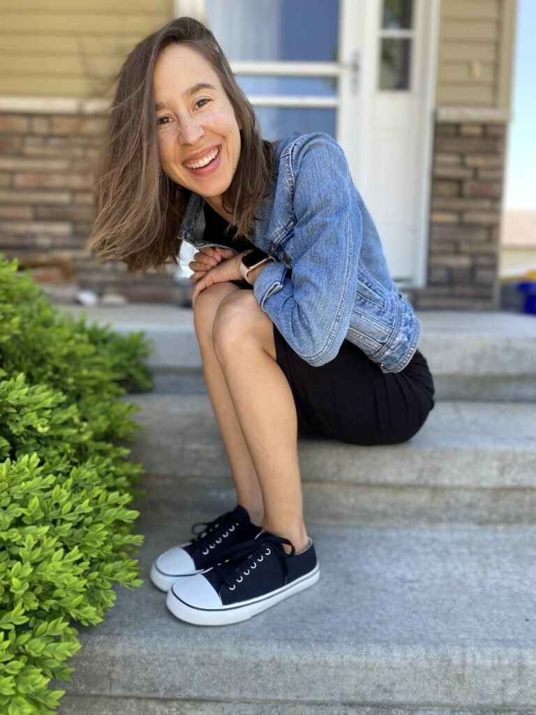 A woman sitting on her front steps in a black dress and jean jacket. She has her arms on her knees and she is smiling at the camera because her feet are happy in her Bohempia Herlik 2.0 hemp barefoot converse dupe sneakers