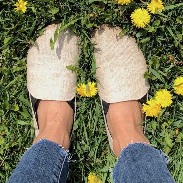 a top down view of a pair of feet standing on green grass wearing Unshoes Terra Vidaa barefot slip-on shoes in natural hemp color