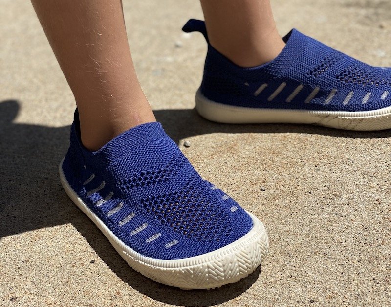 A close up of a child's feet wearing blue Jan and Jul affordable knit slip on shoes standing on concrete