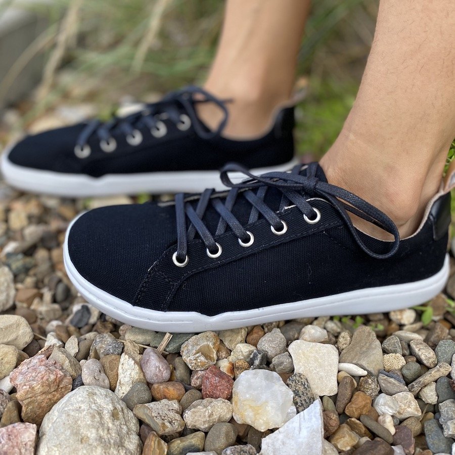 A side view of a pair of feet standing on pebbles wearing Origo Shoes cotton canvas barefoot sneakers in black