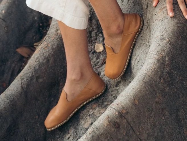 A close up of a pair of feet leaning against a tree trunk wearing Raum goods barefoot slip-on loafers in natural leather