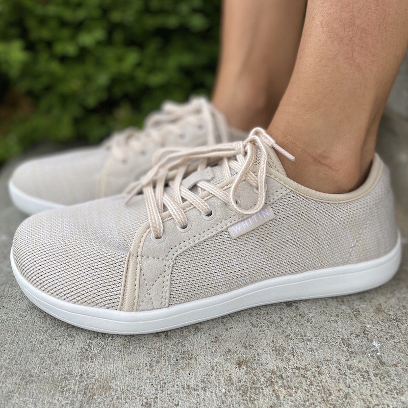 A side view close up of a pair of feet standing on concrete wearing tan Whitin casual barefoot sneakers