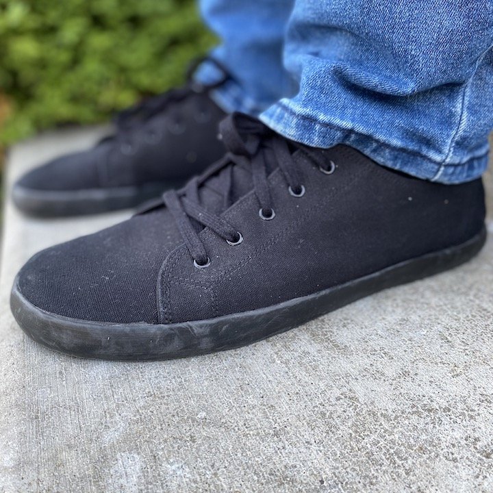 a close up side view of a pair of feet wearing jeans and black Bohempia Hoska barefoot sneakers standing on concrete