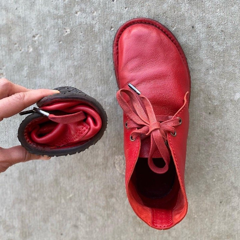 A close up of a pair of Barefoot Pals Gaucho Ninja red leather barefoot desert boots with the left shoe rolled up into a ball to show its extreme flexibility