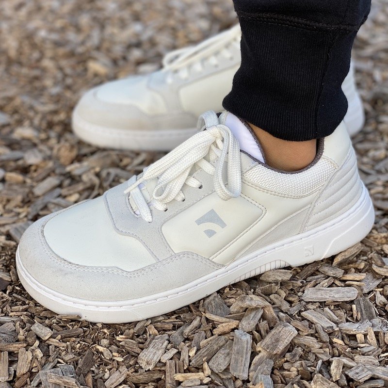 a side view close up of a pair of feet standing on wood chips wearing Barebarics white grey Axiom chunky dad barefoot sneakers