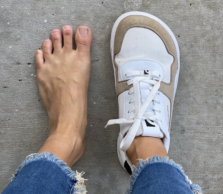 a close up of a pair of feet standing on concrete, the left foot is bare and the other foot is wearing a Barebarics barefoot sneaker, Zing model in taupe, to show it's natural foot shape and wide toe box