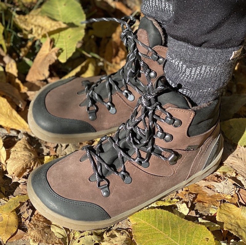 A close up side view of a pair of feet standing on mud and fallen leaves wearing brown Be Lenka Ranger barefoot waterproof hiking boots