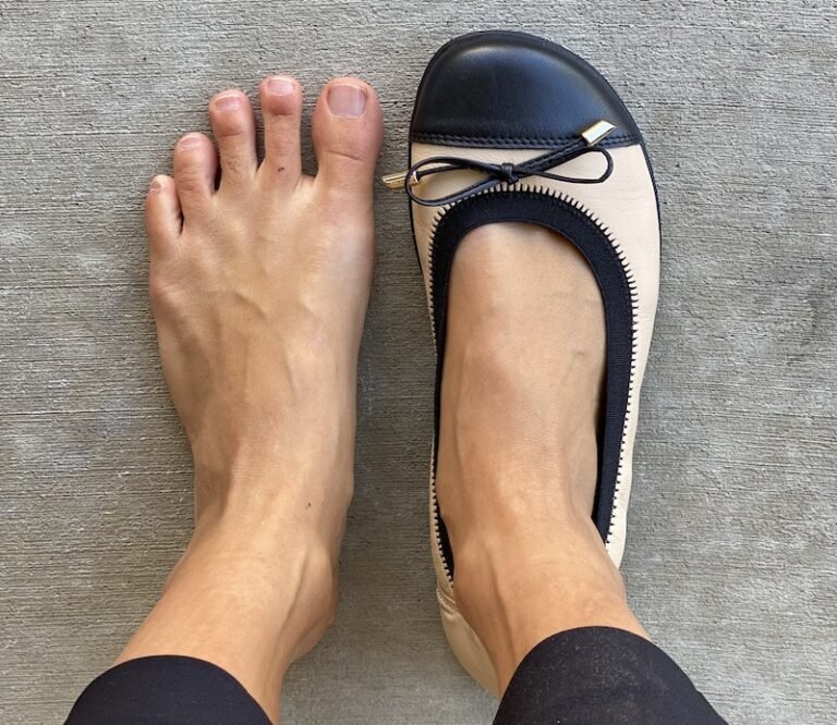 A pair of feet standing on concrete, the left foot is bare and the right is wearing a Groundies Felicia barefoot ballet flat in beige with a black toe cap