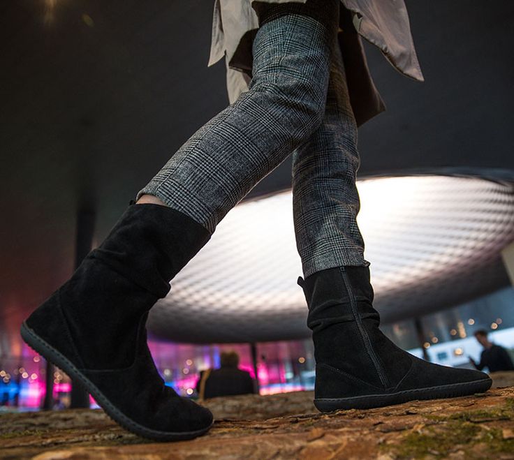 A close up of a person's legs wearing plaid pants and a pair of black suede Groundies Odessa barefoot boots