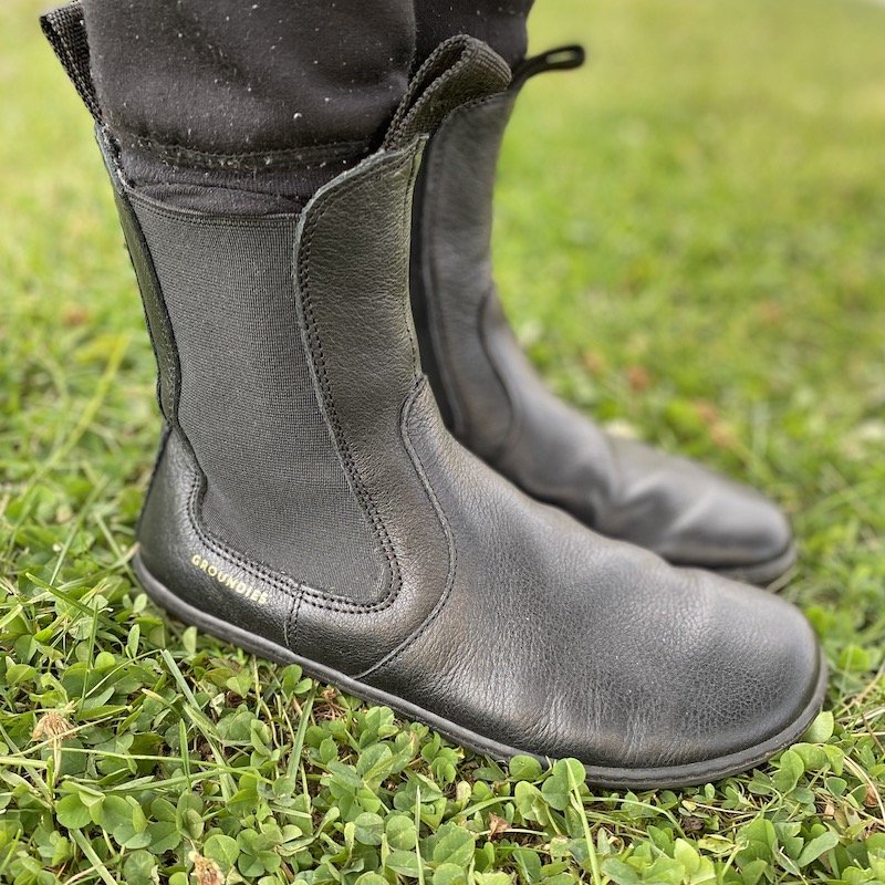 A close up side view of a pair of feet standing on grass wearing Groundies boots, one of the best barefoot boot brands