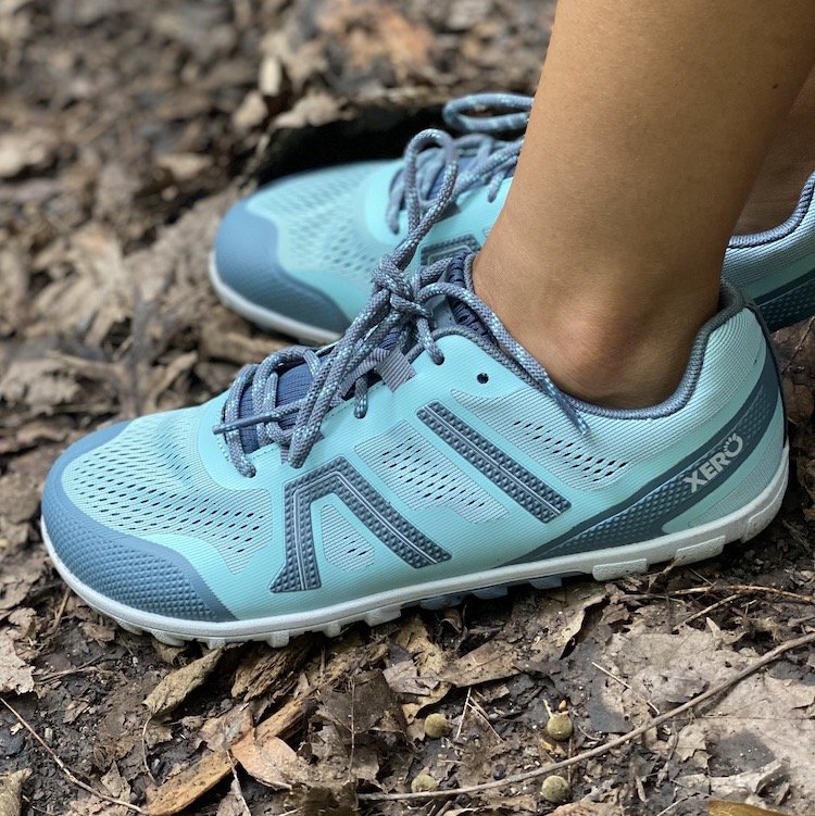 a side view of a pair of feet standing on dirt and leaves wearing Xero Shoes Mesa II lightweight trail hiking barefoot shoe in blue vegan showing the lugs and tread