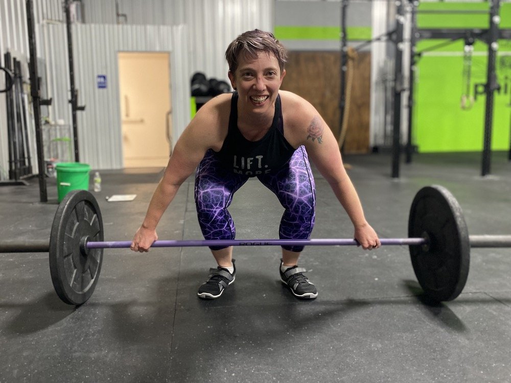 A person in a gym wearing barefoot shoes Xero Forza trainer about to lift a barbell smiling at the camera