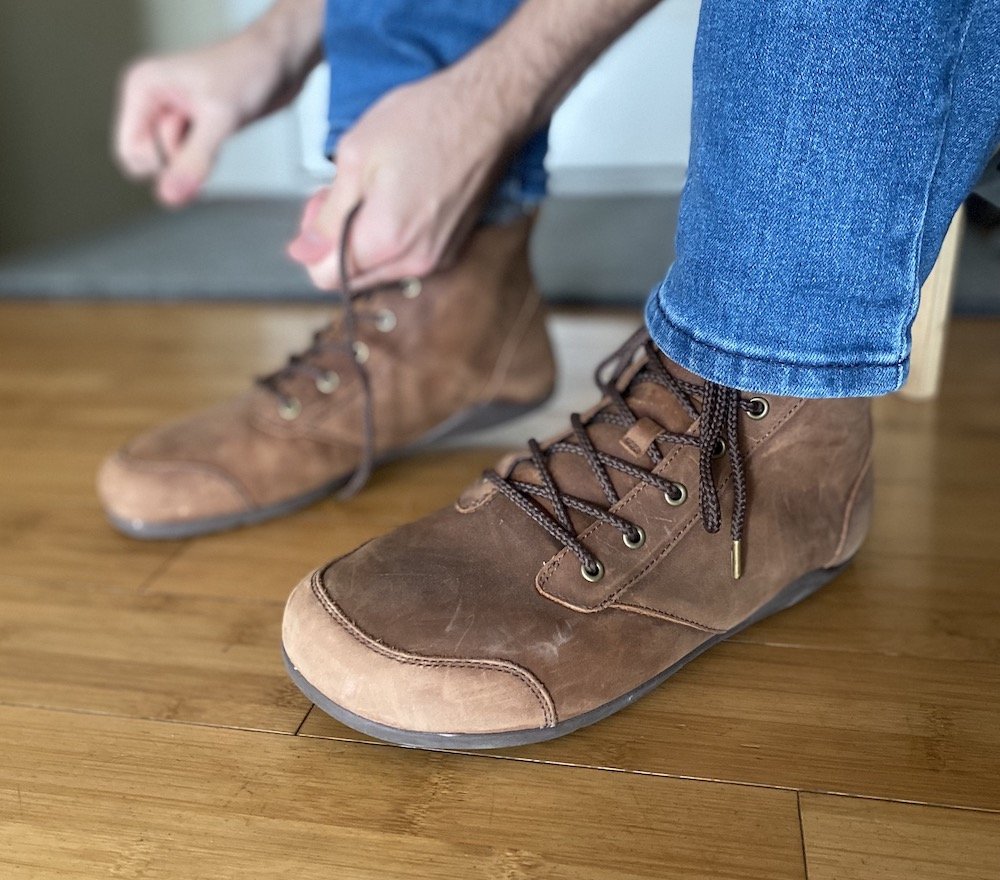 A close up side view of a man putting on a pair of brown leather Denver barefoot boots from Xero Shoes