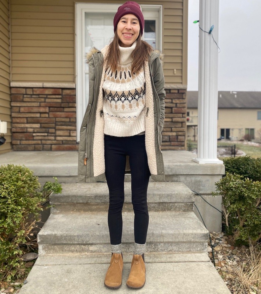 A woman standing on her porch smiling at the camera wearing a sweater, coat, hat, and Lems Chelsea barefoot boots in Cedar