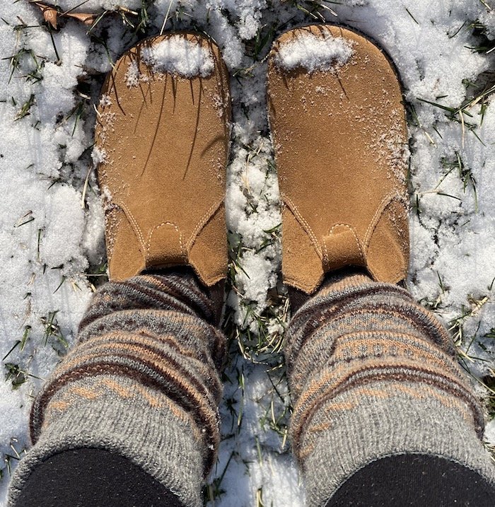 a top down view of a pair of feet standing on snow and grass wearing leg warmers and LEms Chelsea barefoot minimalist zero drop boots in Cedar