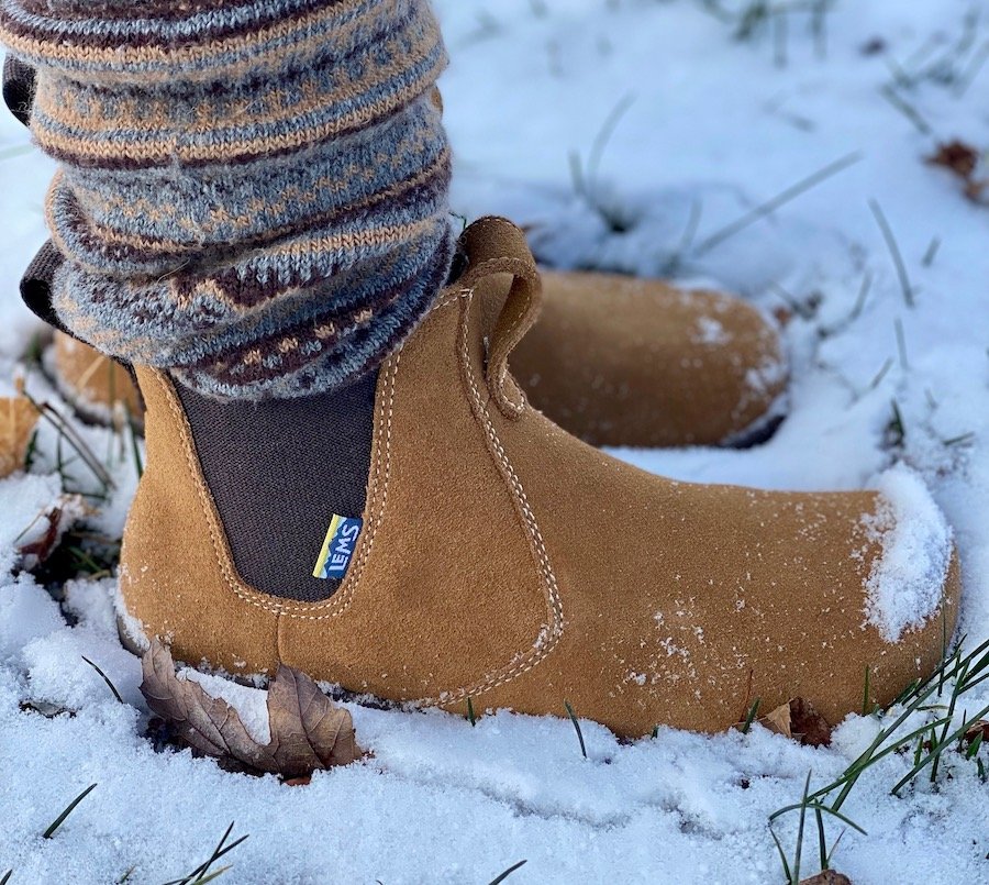 A close up side view of a pair of feet standing in snow wearing leg warmers and Lems Chelsea zero drop boots in Cedar suede leather