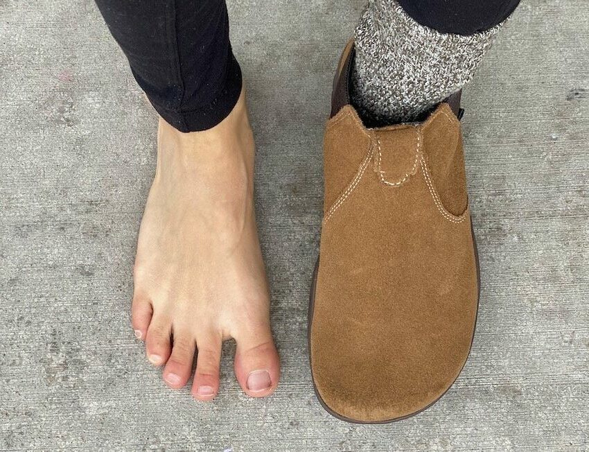 a top down view of a pair of feet standing on concrete. Left foot is wearing Lems Chelsea boots in Cedar, right foot is bare, showing that the boots have an anatomical foot shape