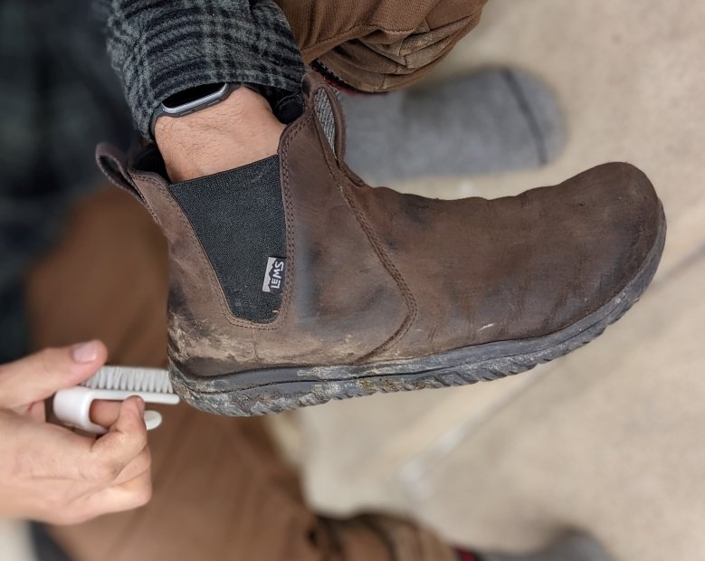 a close up of a hand holding a dirty Lems Chelsea Espresso waterproof minimalist zero drop boot with a shoe brush cleaning off the dirst