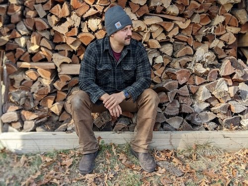 A man sitting in front of a pile of chopped wood wearing work pants, a flannel shirt, and Lems Chelsea Espresso waterproof barefoot boots